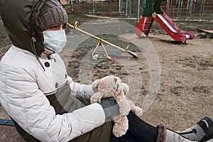 Concept of epidemic and quarantine - a girl with a face mask and a cuddly toy alone on the playground in the city