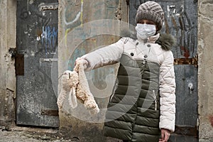 Concept of epidemic and quarantine - a girl with a face mask and a cuddly toy alone on the street in the city