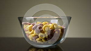 Concept of food and breakfast. Stock footage. Close up of colorful ring corn flakes in a transparent glass bowl on dark