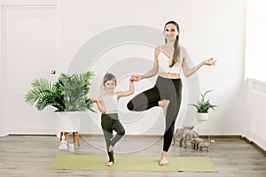 Concept of friendly family and yoga exercises. Pretty young mother and daughter doing yoga Tree pose. Woman and little