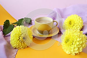 The concept of a good autumn morning. A yellow cup of coffee , yellow dahlia flowers on a lilac light scarf on a multicolored