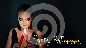 Concept of Halloween. Girl in image of witch with burning candle in hands on black background with inscription Happy