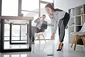 Concept harassment. young woman bend down to take pen from floor. She looks lovely. Her boss sit at table on chair