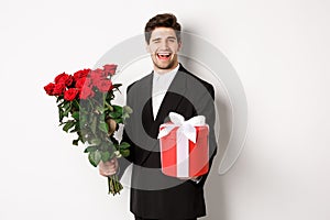 Concept of holidays, relationship and celebration. Charming young man in black suit, holding gift box and bouquet of