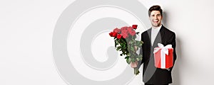 Concept of holidays, relationship and celebration. Charming young man in black suit, holding gift box and bouquet of