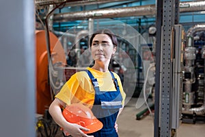 The concept of industrial manufactory. Portrait of a young smiling woman in uniform holds a safety helmet in her hand. Boiler room