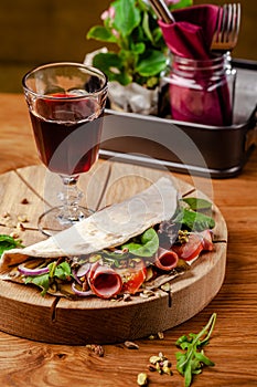 Concept Italian cuisine. Piadina with ham, tomatoes, mix lettuce, pistachios, cucumbers on wooden board. Glass of red wine