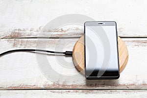 Concept of modern technologies. On the wireless charger is a black smartphone with a glossy white screen. White wooden background