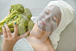 Concept of natural facial skin care. Funny young woman with cosmetic mask made of gray clay, her hair and body wrapped