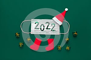 Concept for new year 2022 and christmas. Santa claus hat and felt boots on green background with medical protective mask