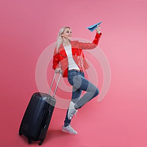 Concept, ready to trip. Young woman with travel suitcase launches paper airplane. Blonde tourist girl on pink background