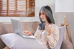 Concept of relaxation at home, Young Asian woman surfing social media on laptop and drinking coffee