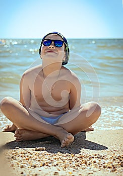 The concept of relaxation and tranquility. A teenager boy sitting on the sand in swimming trunks, blue sun glasses, is engaged in