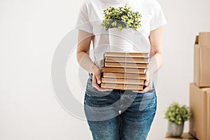 The concept of relocation and moving to a new home. Close-up, female hands hold a pile of books and a green plant in a