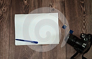 Camera, memory cards, a blank notepad with a pen close-up on a wooden background.