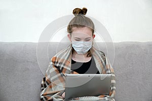 The concept of self-isolation, distance learning. Girl sitting on a sofa in a medical mask wrapped up in a blanket with a laptop