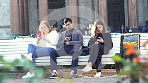 Concept of smartphone addiction. Media. Group of friends, one man and two women sitting outdoor on a bench in park and