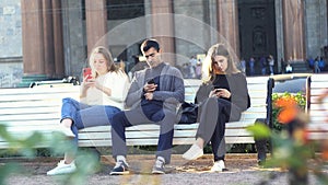Concept of smartphone addiction. Media. Group of friends, one man and two women sitting outdoor on a bench in park and