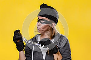 The concept of theft. A woman in a hat, mask and gloves holds the jewels in both hands and examines them. Yellow background and