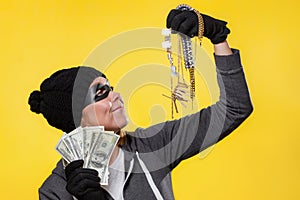 The concept of theft. A woman in a hat, mask and gloves holds a wad of dollars and jewelry in both hands, smiling at them. Yellow