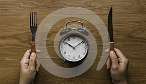 Concept of time and eating with a hand holding fork and knife and a vintage alarm clock on a wooden table