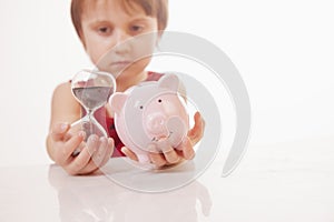 Concept: time is money. Humorous portrait of cute little business child girl holding a sand clock and piggy bank. Selective focus