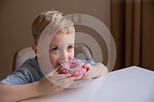 The concept is useless food. A little boy with blond hair eats a pink doughnut at home. He`s happy, he likes it