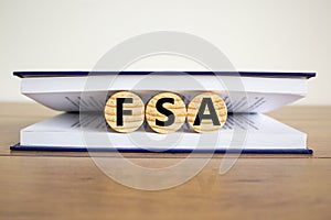 Concept word `FSA, flexible spending account` on wooden circles between pages of a book on a beautiful wooden table. White