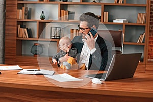 Conception of multitasks. Businessman in formal clothes is working indoors. With toddler