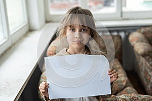 Conceptual image of a sad dejected little girl, holds a blank sheet of paper on gray background.