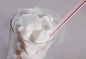 Conceptual macro still life image of refreshment glass full of sugar cubes and straw on white background in glucose addic