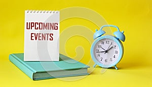 Conceptual message UPCOMING EVENTS in notepad and on yellow background, next to alarm clock and green diary.