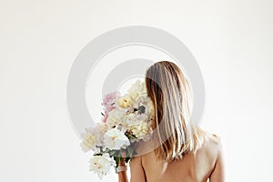 Conceptual portrait of young naked woman with blonde hair from behind with bouquet of white peonies on beige background