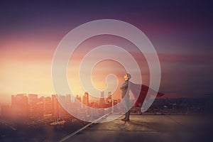 Conceptual sunset scene, business person with red cape stands confident on rooftop looking determined as a superhero over city