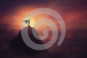 Conceptual sunset scene, superhero with cape standing brave on top of a mountain looks determined at horizon raising one hand up