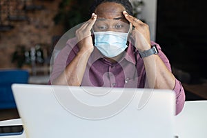 Concerned African-American man wearing protective medical mask staring at the laptop
