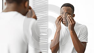 Concerned African Man Touching Bags Under Eyes Standing In Bathroom