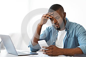 Concerned black freelancer guy looking at smartphone while working at home office