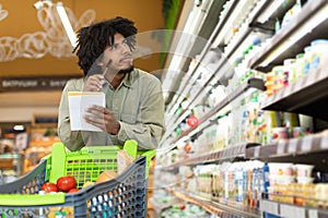 Black Man Holding Shopping List Calculating Food Prices In Supermarket
