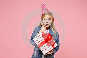 Concerned little kid girl 12-13 years old in denim jacket, birthday hat isolated on pastel pink wall background