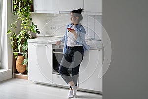 Concerned millennial Asian woman standing in kitchen holding mobile phone waiting for important call