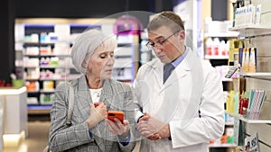 Concerned old female customer asking middle-aged chemist about product reflected on her phone in large pharmacy