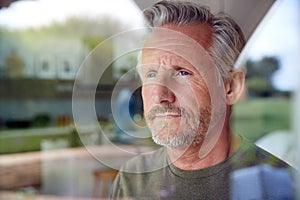 Concerned Senior Man Standing And Looking Out Of Kitchen Door Viewed Through Window