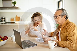 Concerned senior spouses man and woman sitting at table in kitchen in front of laptop, checking bills, making countings