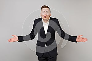 Concerned shocked young business man in classic black suit, shirt shrugging shoulders, spreading hands isolated on grey