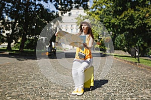 Concerned traveler tourist woman in yellow clothes hat sit on suitcase looking on city map search route in city outdoor