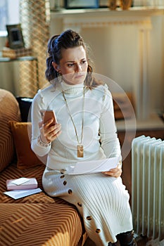 Concerned woman near radiator with smartphone and utility bill
