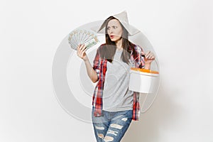 Concerned woman in newspaper hat holding bundle of dollars, cash money and empty paint bucket with copy space isolated