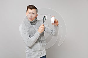 Concerned young man in gray sweater, scarf holding medication tablets, aspirin pills in bottle and magnifying glass