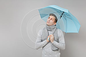Concerned young man in gray sweater, scarf looking aside holding blue umbrella isolated on grey background. Healthy